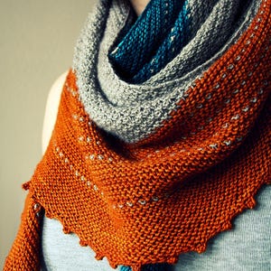 May include: A knitted shawl in shades of orange, grey, and teal. The shawl has a textured appearance with a scalloped edge. The orange section features small white accents. The shawl is draped over a person's shoulders.