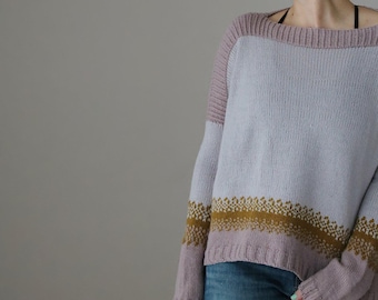 Op de afbeelding: Een lichtblauwe trui met mauve mouwen en halslijn. De onderkant van de trui heeft een band met gouden en mauve geometrische patronen. De trui is gemaakt van zacht gebreid materiaal.