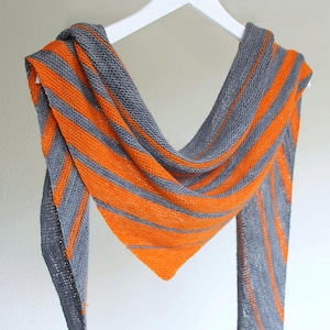Peut inclure: Un châle tricoté à rayures orange et grises. Le châle est suspendu à un cintre blanc, mettant en valeur sa forme triangulaire et son motif tricoté texturé. La combinaison de couleurs crée un contraste visuel saisissant.
