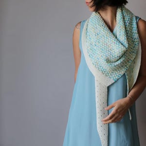 May include: A knitted shawl in shades of blue, green, and cream. The shawl has a textured pattern and a decorative border. The shawl is draped over the shoulders. The background is a solid grey.