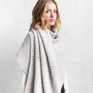 Könnte beinhalten: Ein hellgrauer Strickschal mit strukturiertem Muster und Rollkragen. Der Schal ist über die Schultern drapiert, mit einem Abschnitt mit einer Reihe kleiner, gleichmäßig verteilter Löcher. Das Kleidungsstück ist aus weichem Material, möglicherweise Wolle.