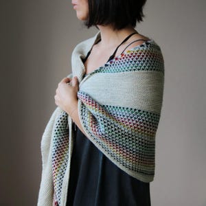 May include: A light beige knitted shawl with a rainbow striped pattern. The shawl is draped over the shoulders. The knitted pattern has a geometric design. The shawl is made of soft yarn.