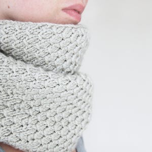 May include: A close-up of a light grey knitted cowl scarf. The scarf has a thick, textured yarn with a basketweave pattern. The cowl is wrapped around the neck, with the lower part of the face visible.