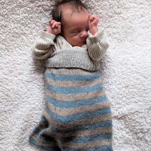 May include: A sleeping baby wearing a light beige sweater and a blue and tan striped sleep sack. The baby is lying on a white, fluffy blanket. The baby's hands are near their face.