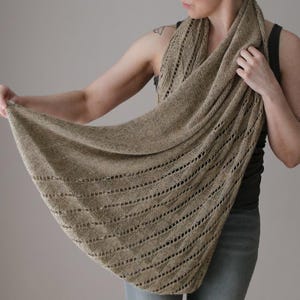 Könnte beinhalten: Ein gestrickter Schal in einem hellbraunen Farbton mit einem strukturierten Lochmuster. Der Schal ist drapiert und hebt seinen fließenden Stoff und sein kompliziertes Muster hervor. Die Kanten des Schals haben eine dekorative Bordüre. Der Schal ist aus weichem Material.