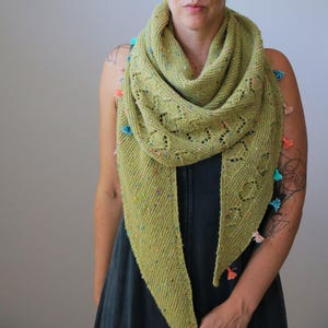 May include: A light green knitted shawl with colourful flecks and decorative openwork. The shawl is adorned with small tassels in shades of pink, blue, and orange. The triangular scarf is draped around the neck.