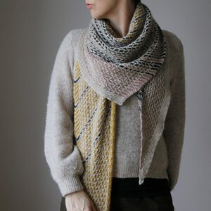 May include: A knitted shawl in shades of beige, pink, yellow, and navy blue. The shawl is draped over a beige sweater. The shawl features a geometric pattern and a textured design. A stylish accessory for cooler weather, perfect for the UK climate.
