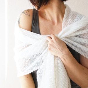 May include: A white, sheer knit shawl draped over the shoulders. The shawl has a delicate, open weave and a soft texture. The garment is lightweight and appears to be made of a natural fiber. The shawl is wrapped around the upper body.