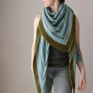 May include: A light blue knitted shawl with a geometric design. The shawl has a dark olive green border with black stripes. The shawl is draped over the shoulders and is made of soft yarn. The shawl is a triangle shape.