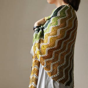 May include: A knitted shawl with a zig-zag pattern. The shawl has a colour gradient from green to yellow and brown, with white edging. The shawl is draped over a person's shoulders.