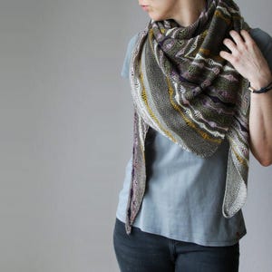 May include: A grey, olive green, and purple knitted shawl with yellow and white accents. The shawl is draped over the shoulders, showcasing its intricate pattern and texture. The shawl is a triangular shape.