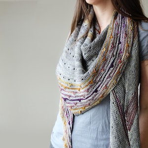 Puede incluir: Un chal de punto gris con un borde colorido. El chal presenta una forma triangular con un patrón de punto texturizado y un borde decorativo en tonos morado, naranja y amarillo. El chal está drapeado sobre los hombros.