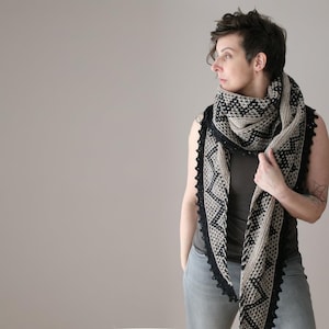 May include: A beige and black patterned shawl with a black lace trim. The shawl is draped around the shoulders and neck. The geometric design is visible on the shawl.