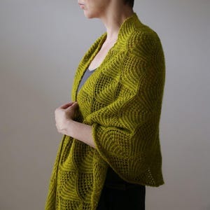 May include: A chartreuse green knitted shawl with a textured, openwork design. The shawl is draped over the shoulders, showcasing the intricate pattern and colour. The garment has a flowing, elegant appearance.