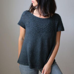 Könnte beinhalten: Ein dunkelblaugrüner Kurzarmpullover mit einem strukturierten Muster auf der Brust. Der Pullover hat einen Rundhalsausschnitt und eine leicht ausgestellte Form. Der Stoff wirkt weich und hat einen dezenten Glanz.