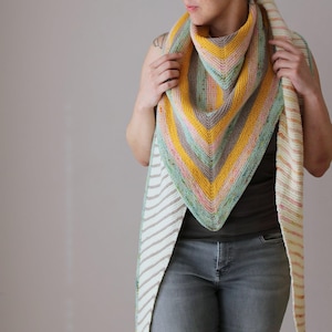 May include: A hand-knitted shawl in a triangular shape, featuring stripes of yellow, pink, green, and grey. The edges have a cream and tan striped pattern. The shawl is designed to be worn around the neck and shoulders.