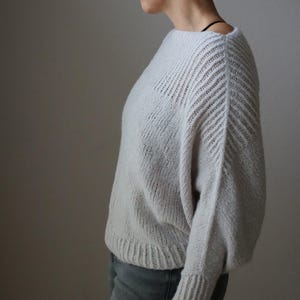 Può includere: Un maglione lavorato a maglia grigio chiaro con scollo a barchetta e maniche lunghe. Le maniche presentano un motivo a coste diagonali e il maglione ha un orlo a coste. Il colore morbido e neutro lo rende un capo versatile.