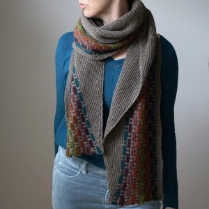 May include: A knitted scarf in grey with a colourful striped pattern. The scarf is wrapped around the neck and drapes down the front. The striped pattern includes colours like orange, green, purple, and blue. The scarf appears to be made of a soft, textured yarn.