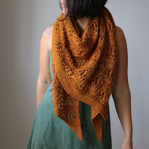 Peut inclure: Un châle tricoté de couleur orange doré. Le châle présente un motif ajouré avec des motifs complexes. La forme triangulaire se drape sur les épaules, créant un accessoire élégant et confortable. Le châle est fait de fil doux.