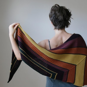 May include: A knitted shawl featuring a striped pattern in autumnal colours: orange, yellow, burgundy, and green. The shawl is held up, displaying its geometric design and textured knit. The colour scheme is warm, suitable for adding a vibrant touch to any ensemble.