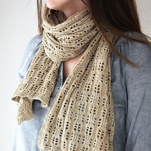 May include: A beige, open-weave scarf is draped around the neck. The scarf has a delicate, lacy pattern with a soft, textured appearance. The scarf is made of a light-coloured yarn, and the design is visible throughout the length of the scarf.