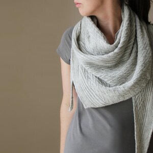 Peut inclure: Un châle tricoté gris clair avec un motif texturé. Le châle est drapé autour du cou et des épaules. Le tissu semble doux et a un éclat subtil. Le châle est de forme triangulaire avec une extrémité pointue.