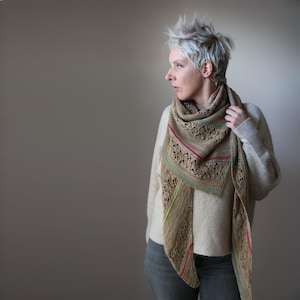 May include: A beige knitted shawl with a lace design and colourful stripes of red, green, and blue. The shawl is draped over a person's shoulders. The person is wearing a cream-coloured jumper.