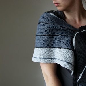 May include: A knitted shawl in shades of grey, black, and white. The shawl has horizontal stripes of varying widths and colours. The fabric appears soft and textured, suitable for cooler weather.