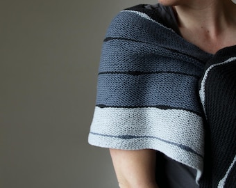 Könnte beinhalten: Ein gestrickter Schal in Grau-, Schwarz- und Weißtönen. Der Schal hat horizontale Streifen unterschiedlicher Breite und Farbe. Der Stoff wirkt weich und strukturiert, geeignet für kühles Wetter.