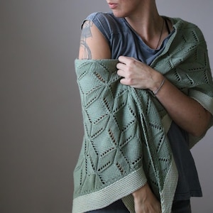 May include: A sage green knitted shawl with a geometric diamond pattern. The shawl has a light green border and is draped over the shoulders. The knitted design features openwork details.