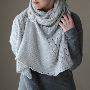 May include: A light gray knitted shawl with a decorative silver brooch. The shawl features a textured design with openwork detailing along the edges. The shawl is draped around the neck and shoulders.