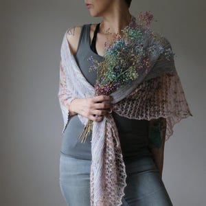 May include: A pale pink and grey knitted shawl with an openwork pattern. The shawl is worn over the shoulders and held with a bouquet of colourful dried flowers. The shawl has a soft, textured appearance.
