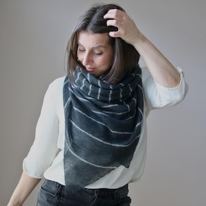 May include: A triangular knit scarf in a dark charcoal gray color featuring thin, horizontal white stripes. The scarf is draped around the neck and shoulders, showcasing a soft, textured fabric with a pointed bottom edge and a wide, folded neckline.