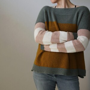 May include: A color-block sweater with a boat neck. The top section is gray, followed by a mustard-yellow band, and the bottom is gray. The sleeves feature alternating stripes of white and pink. The sweater is made of a soft, knitted material.