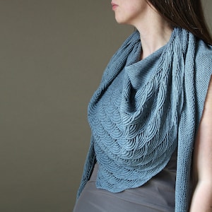 May include: A light blue knitted shawl with a scalloped pattern. The shawl is draped over the shoulders, showcasing the intricate texture and design. The soft blue colour and detailed knitwork create a cosy and elegant accessory.