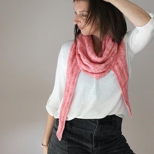 Könnte beinhalten: Ein dreieckiger Strickschal in Korallenrosa mit einem komplexen, durchbrochenen Spitzenmuster. Der Schal ist um den Hals drapiert und zeigt eine weiche, strukturierte Garnkonstruktion mit zarten, sich wiederholenden Ösendetails.