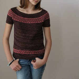 May include: A brown knitted top with short sleeves and a scoop neckline, featuring a pink patterned band at the chest and hem. The top is worn with blue jeans. The knitted texture is visible.
