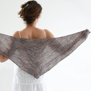 May include: A triangular shawl in shades of gray and brown is held up, showcasing its knitted texture. The shawl appears to be made of a soft, lightweight yarn. The person holding the shawl is wearing a white dress.