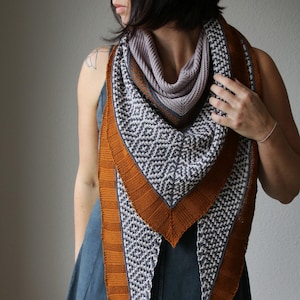 May include: A knitted shawl with a geometric pattern in grey and white, framed by a rust-coloured border. The shawl is draped over the shoulders, showcasing its triangular shape and textured design. The inner layer is a solid light purple.