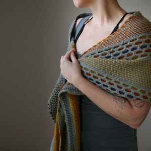 May include: A gray, orange, and navy blue knitted shawl. The shawl features a geometric pattern with openwork details. The color palette includes shades of gray, orange, yellow, and navy blue. The shawl is draped over the shoulders.