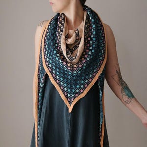 May include: A knitted shawl with a geometric pattern in shades of teal, pink, and peach. The shawl has a peach border and a beige interior. The triangular design is suitable for cool weather and adds a touch of handmade style.