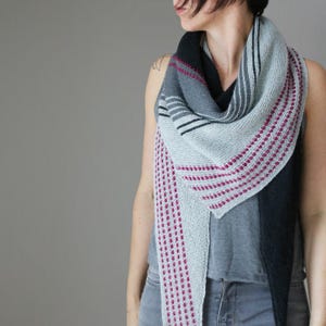 Könnte beinhalten: Ein gestrickter Schal in Grau, Schwarz und Rosa. Der Schal hat ein geometrisches Design mit Streifen und gepunkteten Mustern. Die Farbpalette umfasst Grautöne, Schwarz und Rosa und schafft so ein optisch ansprechendes Accessoire. Der Schal ist um den Hals drapiert.