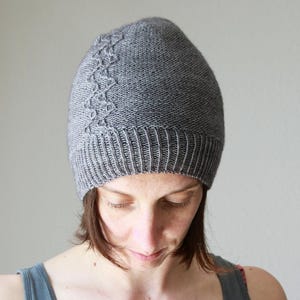 Puede incluir: Un gorro de punto gris con borde acanalado y un diseño de punto de cable decorativo en el lateral. El gorro está hecho de un material suave y cálido, perfecto para el frío. Este gorro hecho a mano es un accesorio elegante.