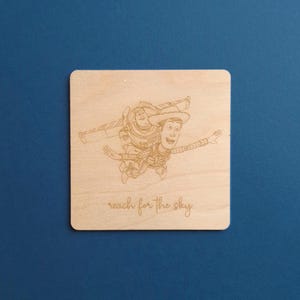 May include: A square wooden plaque with an engraved image of Buzz Lightyear and Woody from Toy Story in mid-air. The phrase "reach for the sky" is etched below the characters. The plaque is made of light-coloured wood.