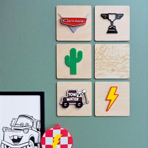 May include: A set of wooden square wall art pieces with diverse designs. Includes a red and silver car logo, a black trophy, a green cactus, a map, a tow truck, and a yellow lightning bolt. The art is displayed on a light green wall.