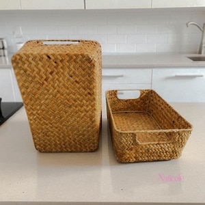 Könnte beinhalten: Zwei gewebte Seegras-Körbe. Einer ist rechteckig und aufrecht mit einem Griffausschnitt. Der andere ist ein rechteckiges Tablett mit einem Griffausschnitt. Beide sind hellbraun und stehen auf einer hellen Arbeitsplatte.
