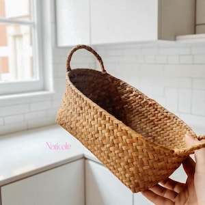 Op de afbeelding: Een rechthoekige gevlochten mand met een handvat, gemaakt van natuurlijk gekleurd stro. De mand, vastgehouden door een hand, heeft een gebogen bovenkant en is leeg. De mand is nieuw en heeft de tekst "Naticole".