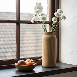 May include: A wooden vase holding white flowers sits on a dark windowsill next to a wooden bowl with fruit. The window frame is dark brown, and the wall is white. The word "Naticole" is in the bottom right corner.