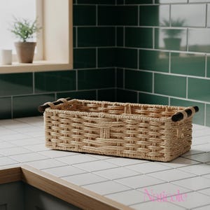 May include: A rectangular woven basket with dark brown handles sits on a white tiled countertop. The basket is made of light brown woven material. The background features a green tiled wall and a window with a small potted plant.