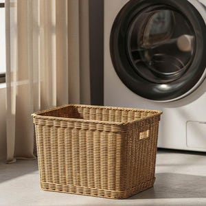 May include: A rectangular, woven wicker laundry basket in a light brown hue. The basket has a square shape with a textured surface and small rectangular handles on the sides. A washing machine is in the background.